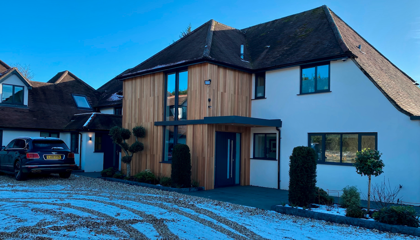 Modern white house with vertical wood cladding and a blue front door; large windows, a black SUV on a snow-dusted gravel driveway under a blue sky.