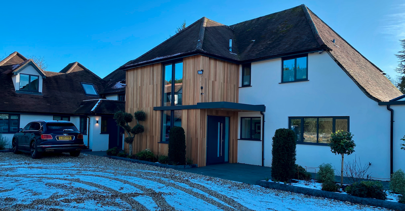 Modern white house with vertical wood cladding and a blue front door; large windows, a black SUV on a snow-dusted gravel driveway under a blue sky.
