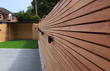 Long wooden fence with horizontal slats beside a paved patio and small patch of green lawn; black wall lights, gravel border, and trees beyond.