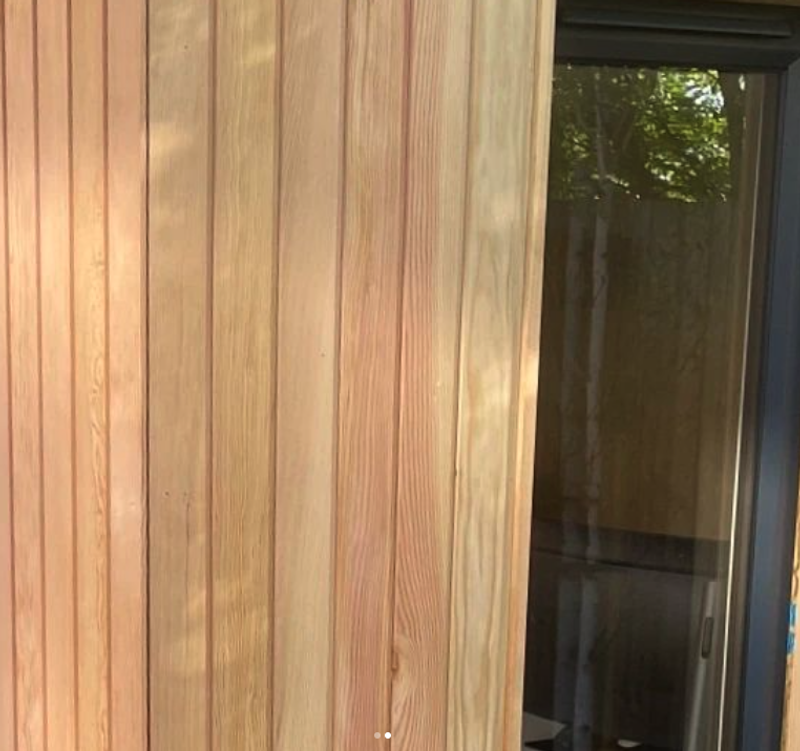 Vertical wooden siding with natural grain on the left and a dark-framed glass door on the right, reflecting outdoor greenery.