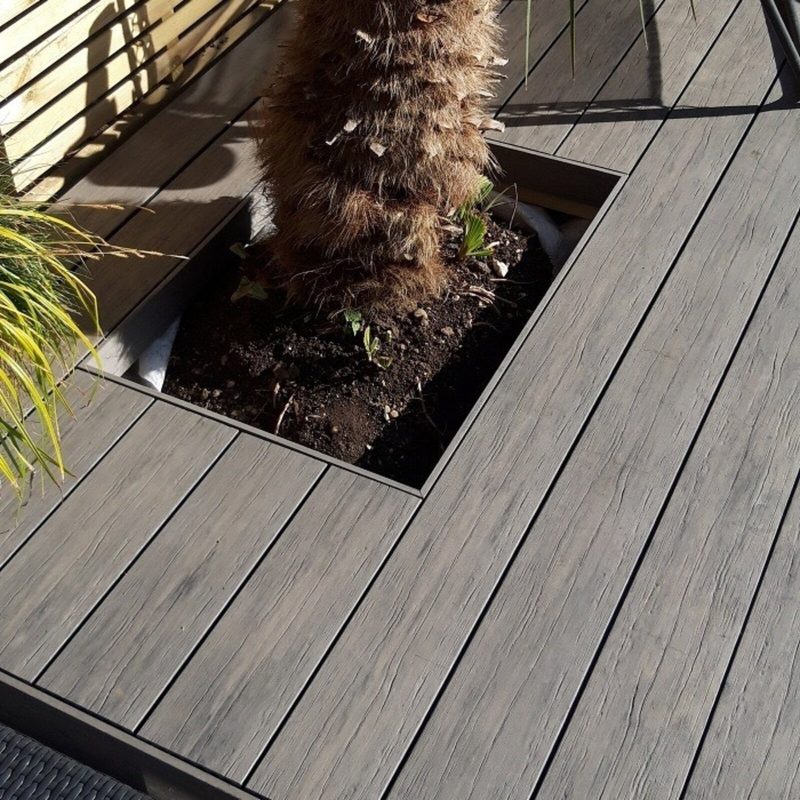 Outdoor deck with a square planter inset containing a palm tree trunk in soil, surrounded by gray wooden planks and some green plants.