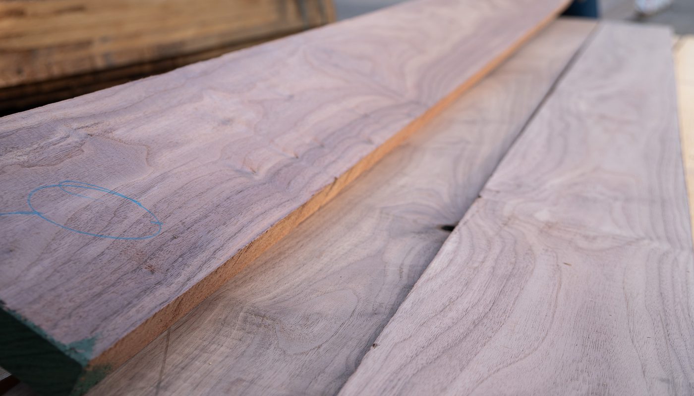 Close-up of long wooden boards stacked outdoors, pale gray-purple grain; blue chalk circle marks one plank, with a green-painted end in the foreground.