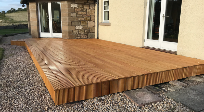 Raised wooden deck with grooved planks beside a beige house, stone wall corner and sliding glass doors; gravel ground and countryside beyond.