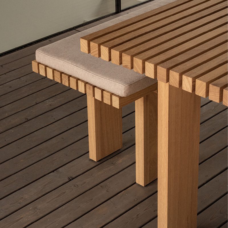 Outdoor wooden table with a slatted top and a matching bench with a beige cushion on a dark wooden deck.