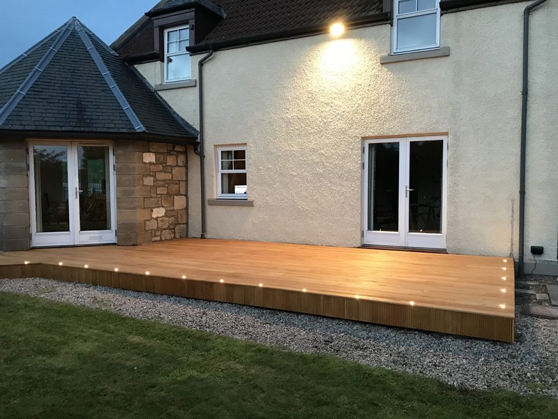 Raised wooden deck along the back of a beige house, with glass double doors, a small window, gravel border, and warm embedded lights at dusk.