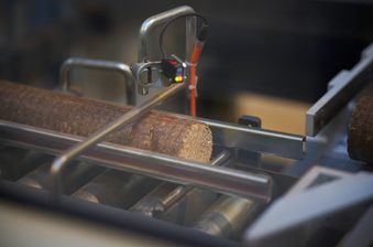 Close-up of a rough log on a metal conveyor in an industrial wood-processing machine, featuring a clamp, a sensor, and orange cables.