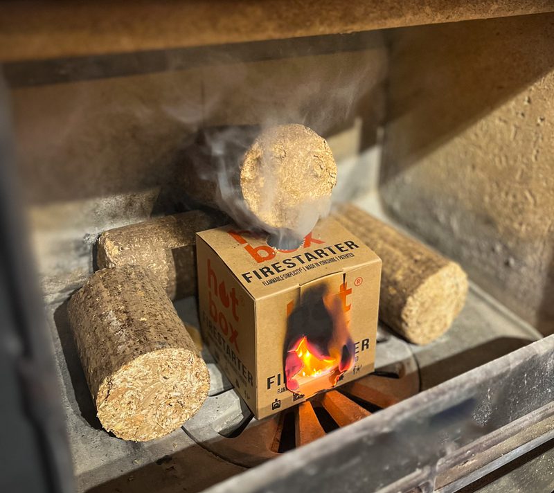 Cardboard firestarter box in a brick fireplace grate, with a round log on top and several logs around; a small flame visible on the box.