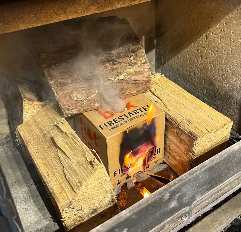 Box labeled FIRE STARTER sits among split logs in a wood-burning fireplace, with flames and smoke rising from the box.