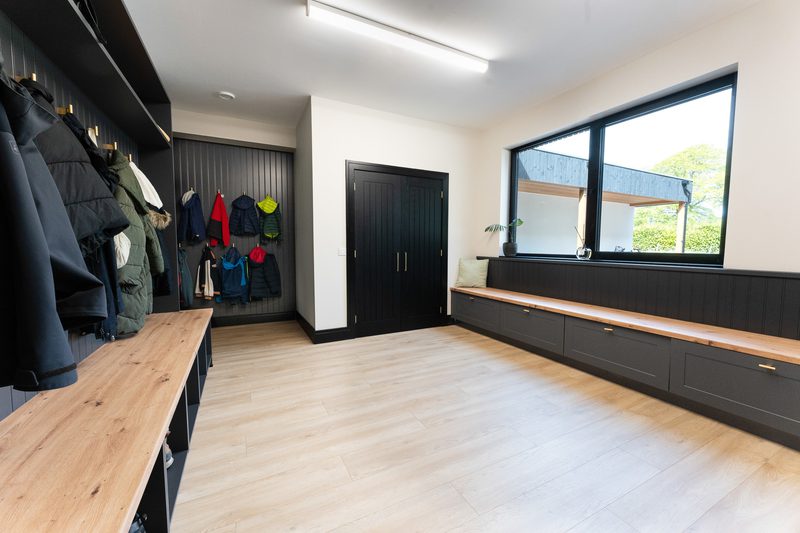 Bright mudroom with light wood floors, dark built-in storage, coats hanging left, long bench with drawers right, a plant on the windowsill, and a large window.