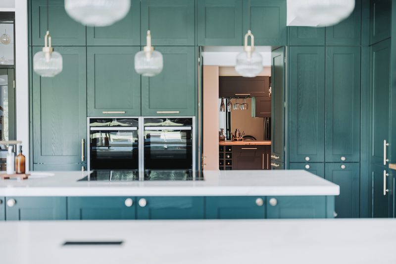 Teal kitchen featuring a white island, built-in double ovens, and globe pendant lights; a doorway leads to a secondary kitchen with dark cabinetry and wine storage.