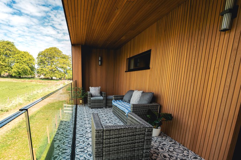 Wood-paneled balcony with wicker seating, grey cushions and a blue throw, patterned tile floor, glass railing, potted plant, overlooking green fields and trees.