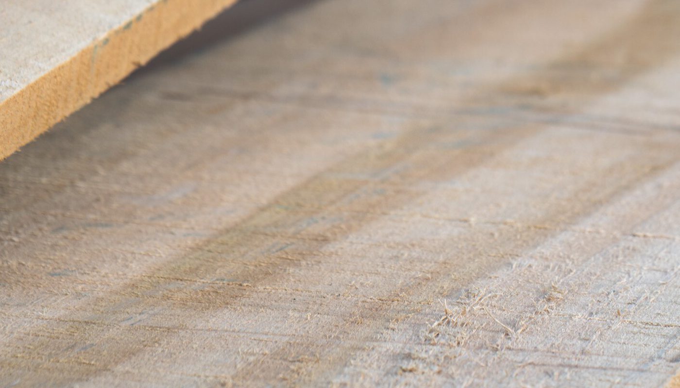 Close-up of a wooden plank resting on a dusty workshop floor, showing grain, rough texture, and scattered sawdust.
