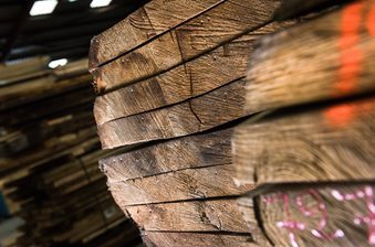 Close-up of rough-cut wooden boards stacked in a warehouse, showing textured ends and grain; a foreground plank has pink chalk markings.