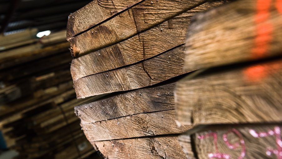 Close-up of rough-cut wooden boards stacked in a warehouse, showing textured ends and grain; a foreground plank has pink chalk markings.