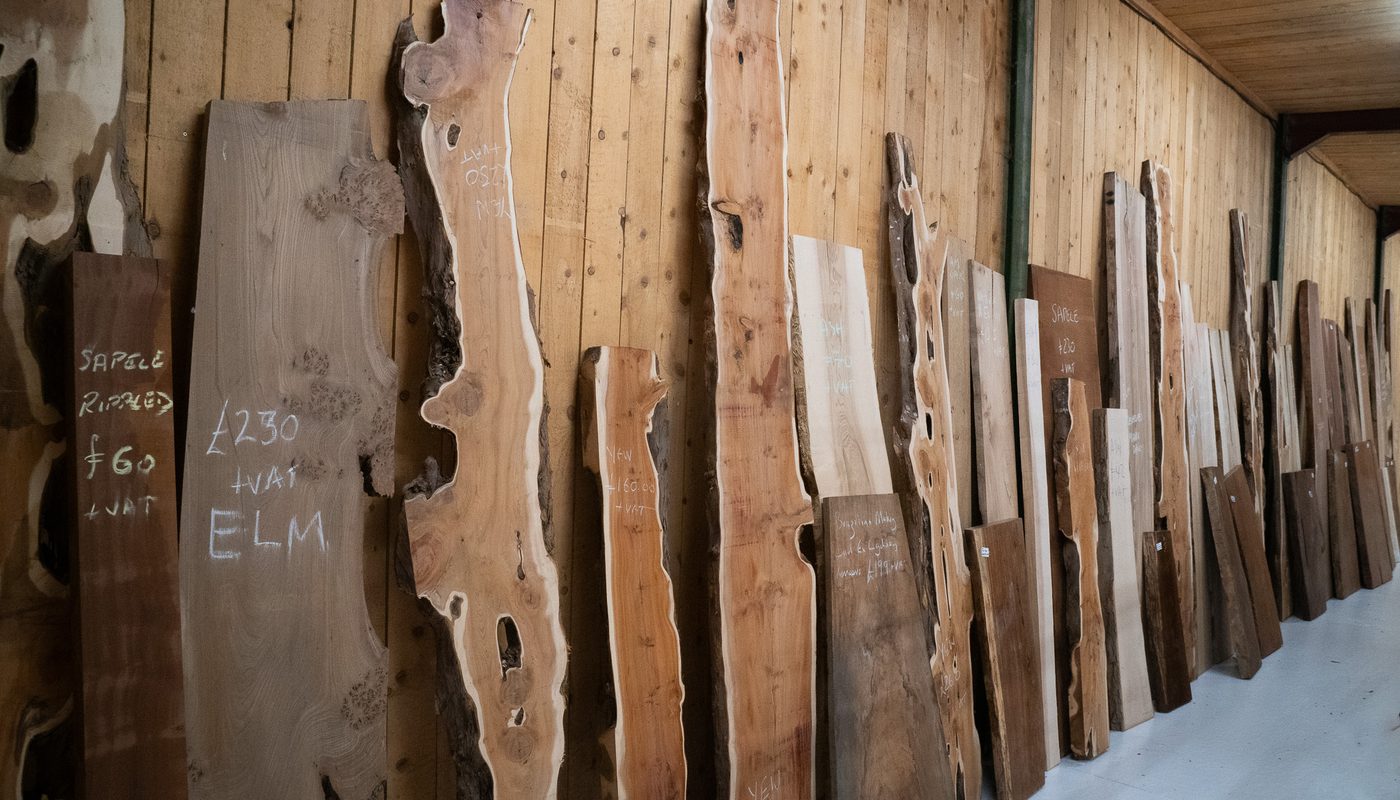 Row of wooden slabs with natural edges leaning against a wood-paneled wall, chalk markings visible on many boards in a lumber storage area.