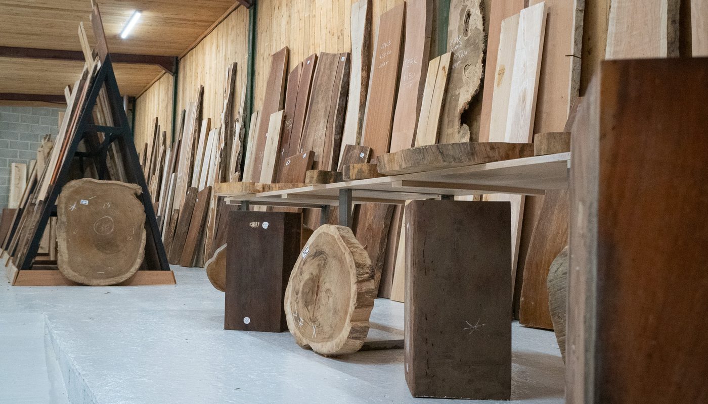 Timber workshop with vertical boards and round wooden slabs lined along the wall, a long bench, and a ladder-like frame on the left.