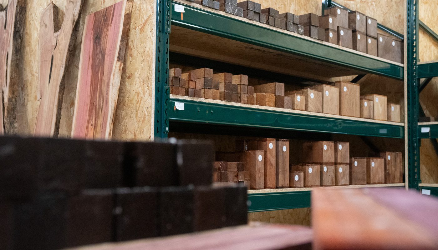 Green metal shelving in a woodshop, neatly stacked wooden blocks and planks of various sizes; plywood sheets lean on the left, foreground blocks blurred.