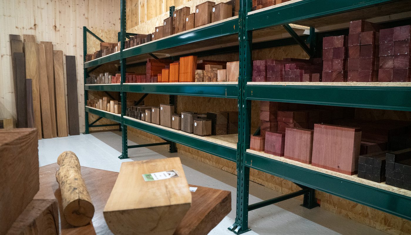 Woodworking shop with green metal shelves holding assorted wooden blocks and planks; a log and a wooden block rest on a table in the foreground.