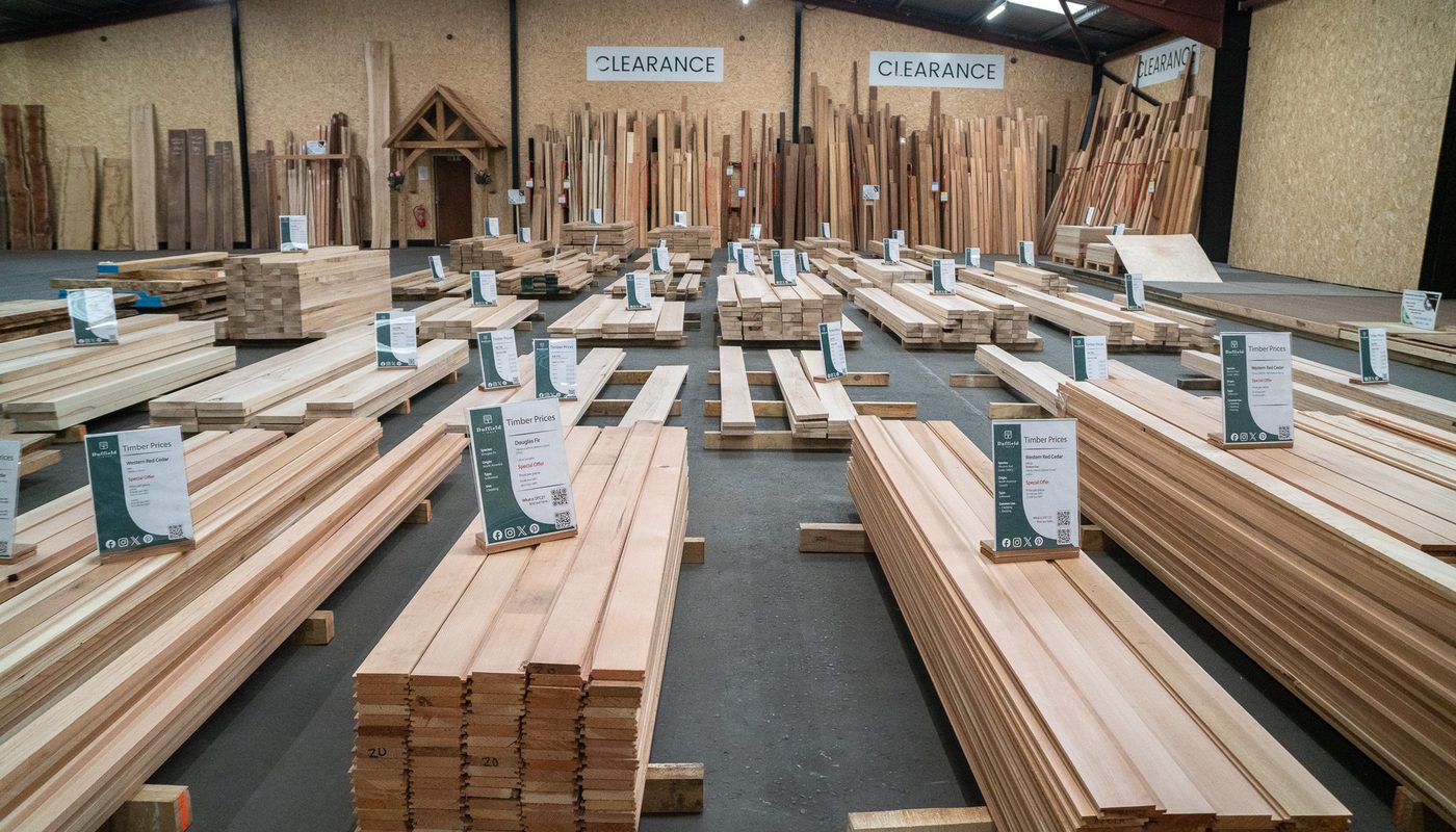 Indoor lumber warehouse with rows of stacked wooden planks and boards on pallets, several sizes; clearance signs line the back wall.