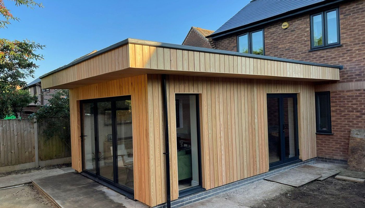 A vertical wooden-panel extension with a flat roof attached to a brick house, featuring large glass doors and a concrete patio in the backyard.