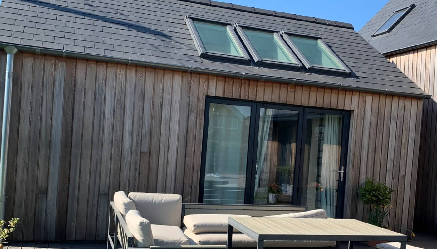 Modern wooden-clad house with a slate roof, three skylights, glass doors, and beige outdoor seating around a rectangular table on a wooden deck.