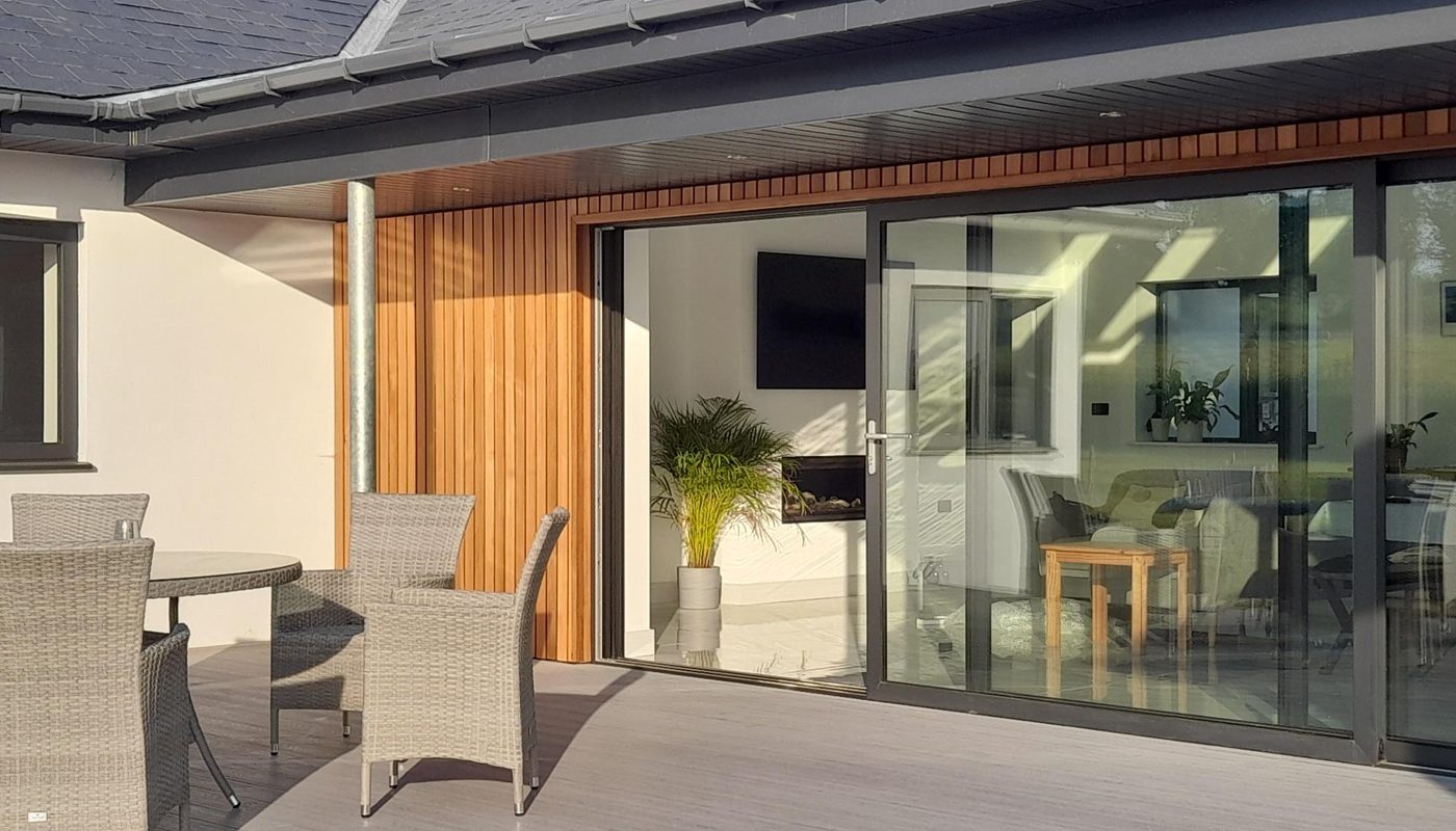Modern home patio with gray decking, a round wicker table and chairs, wooden wall panels, and large sliding glass doors into a bright living area with plants.