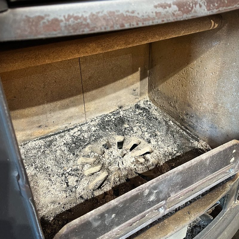 Inside a dusty fireplace insert: ash-covered floor, a circular grate with radial slots, and rusted metal trim around the opening.