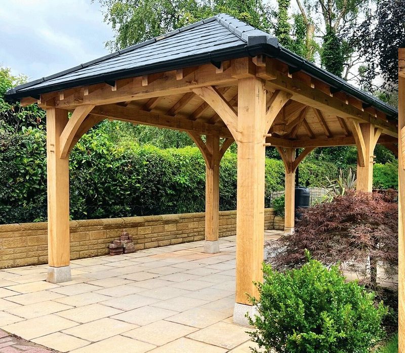 Wooden garden gazebo with tiled roof, sturdy posts, and exposed beams on a paved patio, surrounded by hedges and shrubs.