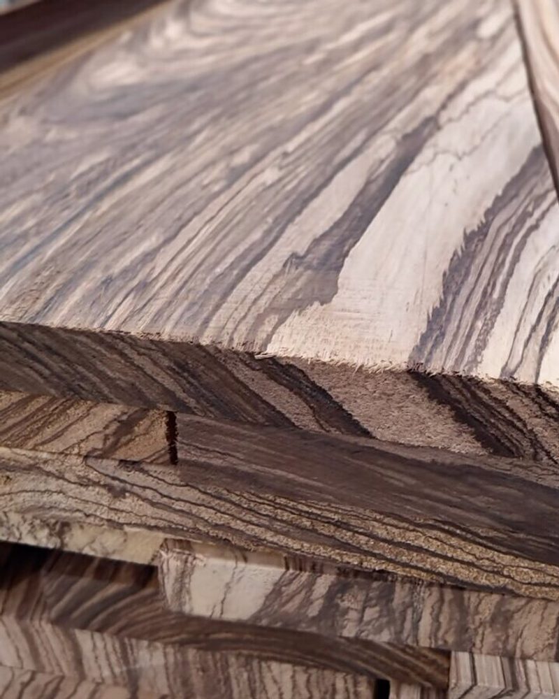 Stack of zebrawood boards with bold dark and light grain, edges visible, resting on a workshop table with blurred tools and shelves in the background.