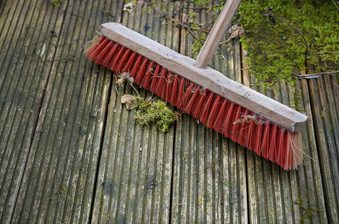 Red-bristled broom with a wooden handle resting on weathered wooden decking, with moss, leaves and small twigs scattered around.