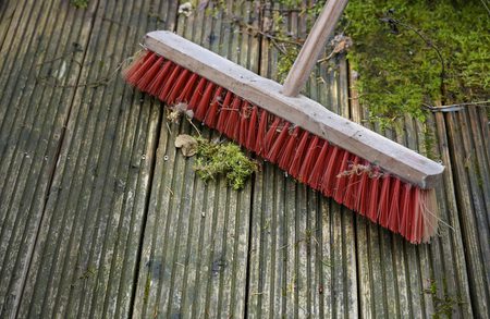 Red-bristled broom with a wooden handle resting on weathered wooden decking, with moss, leaves and small twigs scattered around.
