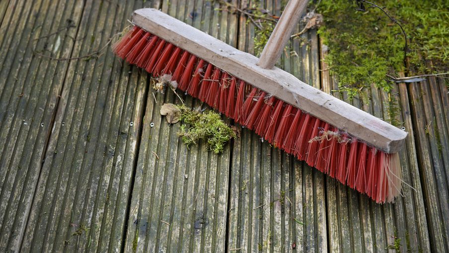 Red-bristled broom with a wooden handle resting on weathered wooden decking, with moss, leaves and small twigs scattered around.