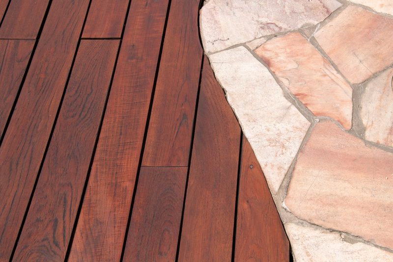 Curved reddish-brown wooden deck boards transition to a pale stone patio with irregular pink-tan stones.