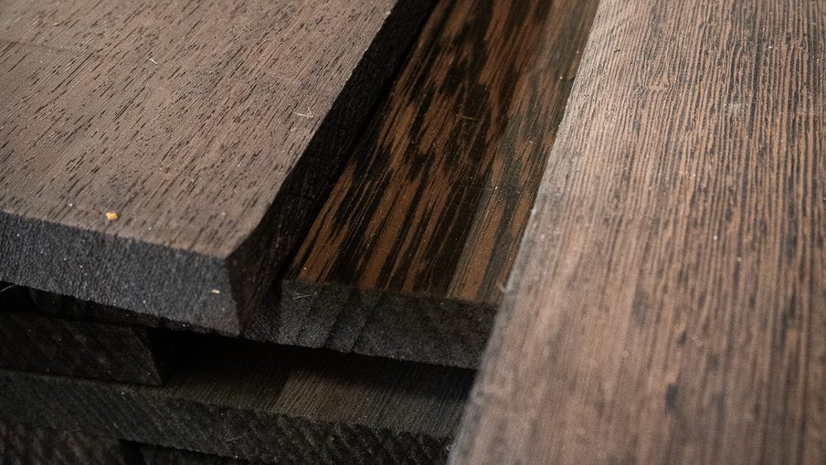 Close-up of dark-stained wooden boards stacked together, showing distinct grain patterns, beveled edges, and layered thicknesses.