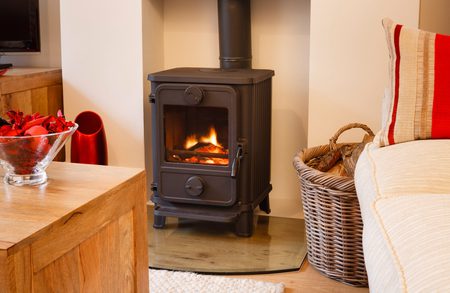 Black wood stove with a glowing fire sits on a glass hearth; a wicker log basket, a red-striped cushion, and a glass bowl of red decor nearby.