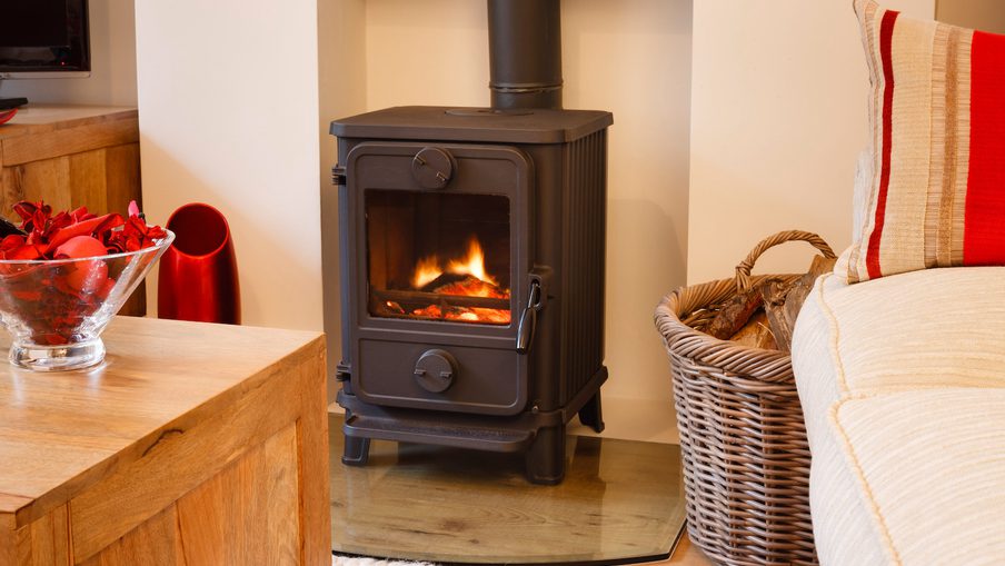 Black wood stove with a glowing fire sits on a glass hearth; a wicker log basket, a red-striped cushion, and a glass bowl of red decor nearby.