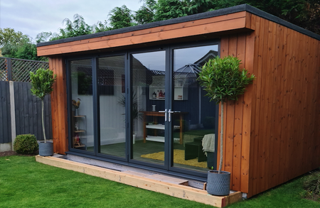Wood-clad garden room with large glass doors and dark frames, on a wooden deck; two potted topiary trees flank the entrance, green lawn beyond.