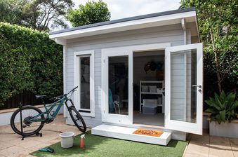 Small gray shed with white trim and open glass doors; a teal mountain bike leaning outside, with a doormat, bucket, spray bottle, and green turf on the patio.
