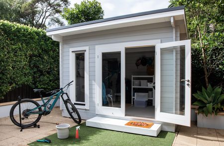 Small gray shed with white trim and open glass doors; a teal mountain bike leaning outside, with a doormat, bucket, spray bottle, and green turf on the patio.