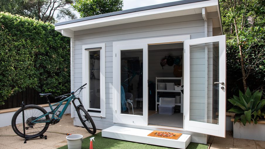 Small gray shed with white trim and open glass doors; a teal mountain bike leaning outside, with a doormat, bucket, spray bottle, and green turf on the patio.
