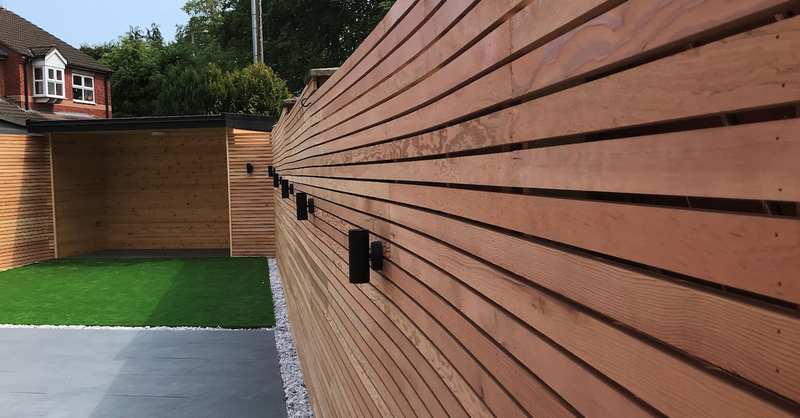 Horizontal wooden privacy fence with black outdoor lights along a modern backyard, a small wooden shed at the far end, green artificial turf, and a gray patio.