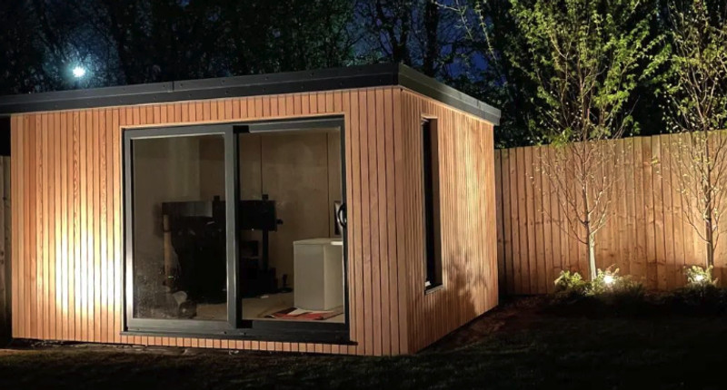 Night view of a small wooden shed with vertical slats and a glass sliding door; interior desk and chair visible, lit trees along a fence.