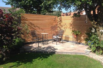 Sunny backyard patio with a wooden slat fence, metal chairs around a small table on a tiled raised deck, and potted plants beside a green lawn.