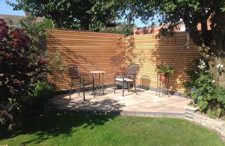 Sunny backyard patio with a wooden slat fence, metal chairs around a small table on a tiled raised deck, and potted plants beside a green lawn.