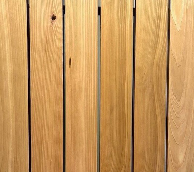 Close-up of vertical wooden planks with visible grain and knots, warm honey-brown color, and narrow gaps between boards.