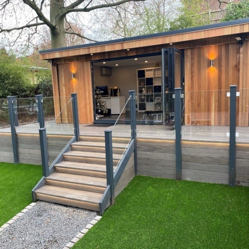 Wooden cabin with open glass doors and interior shelving; a raised deck with glass railing and wooden steps leads to a gravel path and green lawn.