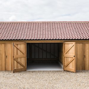 Wooden shed with a red-tiled roof, wide-open central doorway, side doors closed, natural wood panels and black hardware, on gravel under a cloudy sky.