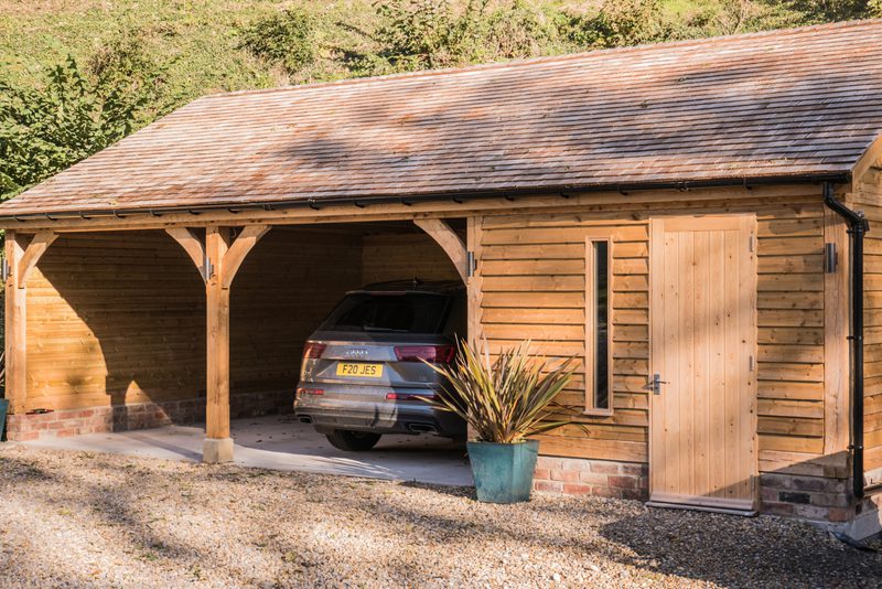 Wooden carport attached to a cabin, with a silver SUV parked inside; curved posts, a wooden door, a narrow window, and a potted plant near the entrance.