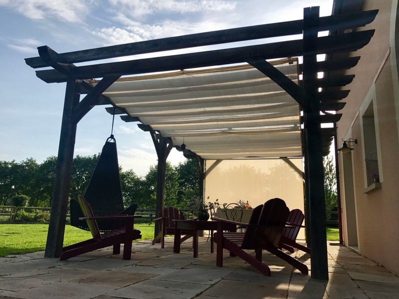Patio under a dark wooden pergola with a white shade; maroon Adirondack chairs around a table and a hanging swing, on a stone patio with green lawn beyond.