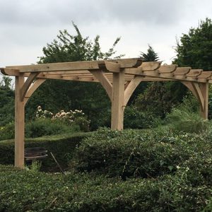 A wooden pergola with curved braces over a hedge-lined garden, featuring flowering bushes and trees beyond under a cloudy sky.